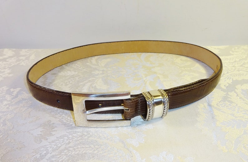 Może przedstawiać: Brązowy sk&oacute;rzany pasek ze srebrną klamrą. Pasek ma teksturowaną powierzchnię i jasnobrązowe wnętrze. Klamra jest prostokątna ze srebrnym wykończeniem. Klasyczny dodatek.