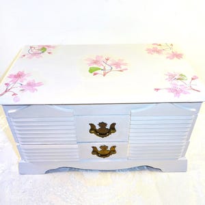 May include: A white wooden jewelry box with two drawers and gold-colored handles. The top of the box is decorated with pink cherry blossom floral designs. The box has a slatted front design.