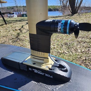 May include: A black and blue Foil Drive MAX electric hydrofoil motor attached to a black and grey hydrofoil board. The motor has a propeller and the text "FOIL DRIVE" in blue. The board is on a grassy surface.