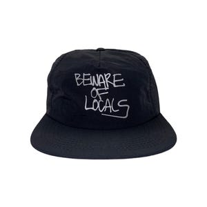 May include: Black cap with a curved peak. The cap has the white text "BEWARE OF LOCALS" in a handwritten style across the front. The cap is made of a smooth material, possibly nylon.