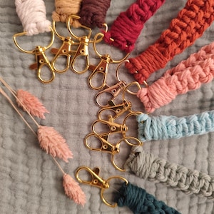 Könnte beinhalten: Eine Sammlung von Makramee-Schlüsselanhängern in verschiedenen Farben, darunter Weiß, Beige, Braun, Rot, Orange, Rosa, Blau, Grau und Türkis. Jeder Schlüsselanhänger hat einen goldfarbenen Verschluss und Ring. Die Schlüsselanhänger liegen auf einem strukturierten, hellblauen Hintergrund.