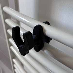 Peut inclure: Un crochet en plastique noir conçu pour se fixer sur une barre de radiateur sèche-serviettes blanc. Le crochet présente une tige en forme de U s'étendant vers l'extérieur depuis une base plate ovale qui maintient l'accessoire sur le rail.