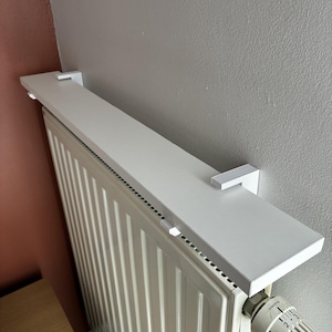 Può includere: Una mensola rettangolare bianca per termosifone montata sopra un radiatore a coste verticali. La mensola presenta due supporti bianchi a forma di L che la fissano all'unità. È visibile una valvola termostatica bianca con impostazioni numerate.