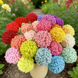 May include: A vase of vibrant, crocheted flowers in shades of red, pink, yellow, green, blue, and purple. The flowers are displayed in a cream-coloured vase. The background shows a blurred garden scene with a stone path.