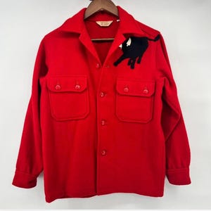 May include: A bright red wool jacket with a black bull applique on the upper left chest. The jacket has two front pockets, a collared neckline, and button closures. The jacket is displayed on a wooden hanger.