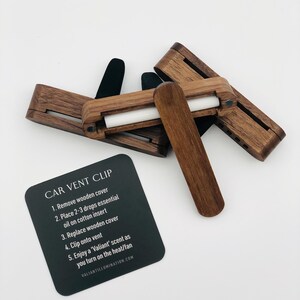 May include: Wooden car vent clips with a dark finish, featuring a white cotton insert. A black card displays instructions: "CAR VENT CLIP" and steps for use. The product is designed to diffuse essential oils in a vehicle.