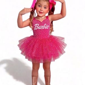 May include: A young girl in a pink Barbie costume, including a sparkly bodice with the Barbie logo and a layered tulle skirt. She wears matching pink bows in her hair and bronze boots.