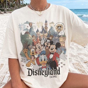 May include: A cream-colored short-sleeve t-shirt featuring a graphic print of Mickey Mouse, Minnie Mouse, Donald Duck, Goofy, and Pluto in front of a castle. The text "Disneyland Est. 1955" is printed in black at the bottom of the graphic design.