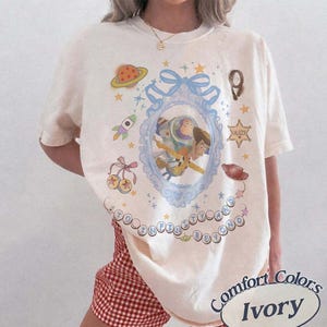 Peut inclure: Un t-shirt ivoire arborant un motif central de Buzz l'&Eacute;clair et Woody dans un cadre orn&eacute; bleu clair avec un n&oelig;ud. Le design comprend une plan&egrave;te, une fus&eacute;e, une &eacute;toile de sh&eacute;rif, un chapeau de cow-boy et le texte TO INFINITY AND BEYOND en bulles.