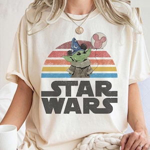 May include: Cream-colored t-shirt featuring a graphic of a green alien character wearing a wizard hat and holding a Mickey Mouse balloon. The design is set against a retro sunset with the words "STAR WARS" in bold, gray letters.