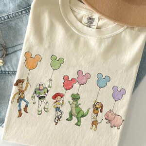 K&ouml;nnte beinhalten: Cremefarbenes T-Shirt mit einer farbenfrohen Illustration von Toy Story-Charakteren, die Mickey Mouse-f&ouml;rmige Ballons halten. Das Design umfasst Woody, Buzz Lightyear, Jessie, Rex, Slinky Dog und Hamm. Das Shirt liegt auf einer Jeans.