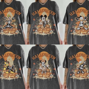 Puede incluir: Una camiseta gris carbón con un estampado gráfico de varios personajes de dibujos animados vestidos con disfraces de esqueleto sentados sobre una pila de calabazas. El diseño incluye la palabra HALLOWEEN en naranja sobre una luna grande.