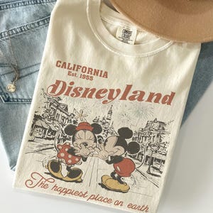 May include: Cream-colored t-shirt with a Disneyland graphic featuring Mickey and Minnie Mouse. The shirt has the text "CALIFORNIA Disneyland Est. 1955" and "The happiest place on earth". The shirt is folded on top of blue jeans.