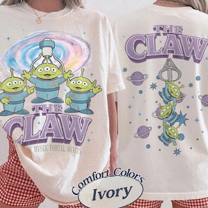 May include: An ivory t-shirt featuring three green three-eyed aliens in blue space suits. The front displays a colorful portal and the text THE CLAW THE MYSTIC PORTAL AWAITS. The back shows the aliens hanging from a claw with the text THE CLAW.
