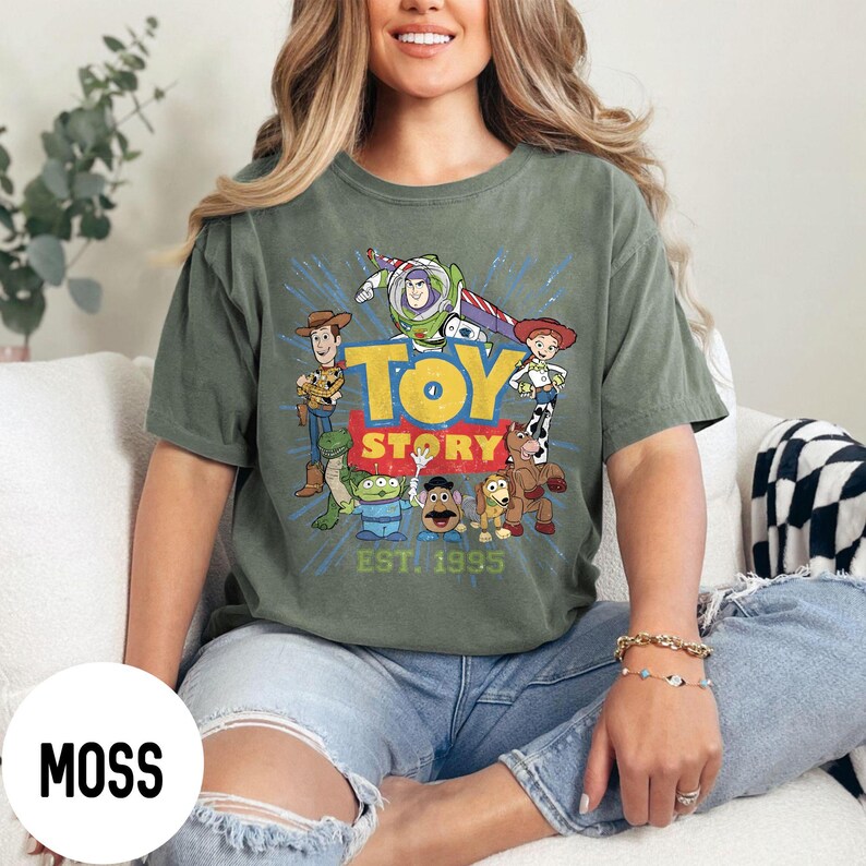 以下が含まれることがあります： モスグリーンのTシャツ。カラフルなトイ・ストーリーのキャラクターのグラフィックと「TOY STORY EST. 1995」の文字がプリントされています。デザインには、ウッディ、バズ・ライトイヤー、その他アニメ映画のキャラクターが含まれています。ゆったりとしたフィット感のシャツです。