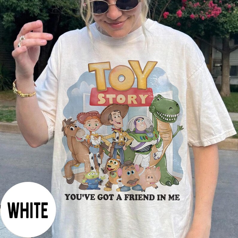 K&ouml;nnte beinhalten: Wei&szlig;es T-Shirt mit einem farbenfrohen Grafikdesign von Charakteren aus dem Animationsfilm "Toy Story". Das Design zeigt Woody, Buzz Lightyear und andere Charaktere mit dem Text "TOY STORY" und "YOU'VE GOT A FRIEND IN ME."