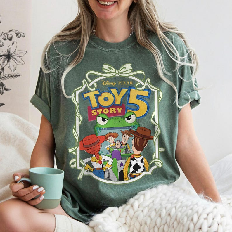 May include: Green t-shirt featuring a Disney Pixar "Toy Story 5" graphic. The design includes characters like Woody, Buzz Lightyear, and Jessie. The shirt has a vintage look and a crew neck.