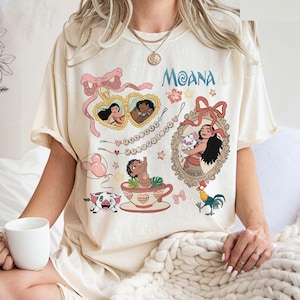 Puede incluir: Camiseta de color crema con un dise&ntilde;o gr&aacute;fico colorido con la palabra "Moana" en azul. El dise&ntilde;o incluye im&aacute;genes de dibujos animados de personajes, una taza de t&eacute;, un cerdo y un gallo. La camiseta tiene mangas cortas.