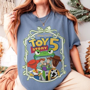 May include: A light blue t-shirt featuring a graphic from the Disney-Pixar movie Toy Story 5. The graphic includes the movie title, a green frog, and several characters, including Woody and Buzz Lightyear. The design is framed with a white border.