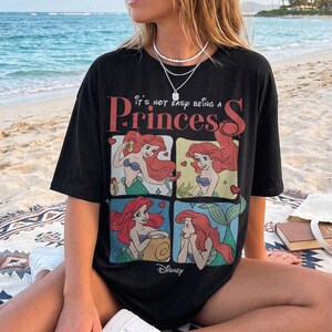 Puede incluir: Una camiseta negra de manga corta con un estampado gr&aacute;fico del personaje Ariel de La Sirenita en cuatro paneles distintos. El texto "It's not easy being a Princess" est&aacute; impreso en rojo sobre los paneles, con el logotipo de Disney debajo.