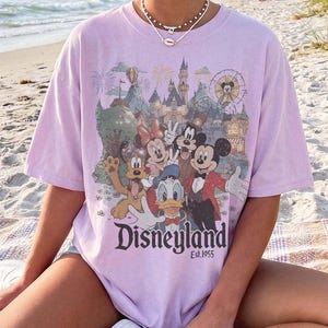 May include: A light purple short-sleeve t-shirt featuring a graphic print of Mickey Mouse, Minnie Mouse, Donald Duck, Goofy, and Pluto in front of a castle. The text Disneyland Est. 1955 is printed below the characters in a dark serif font.