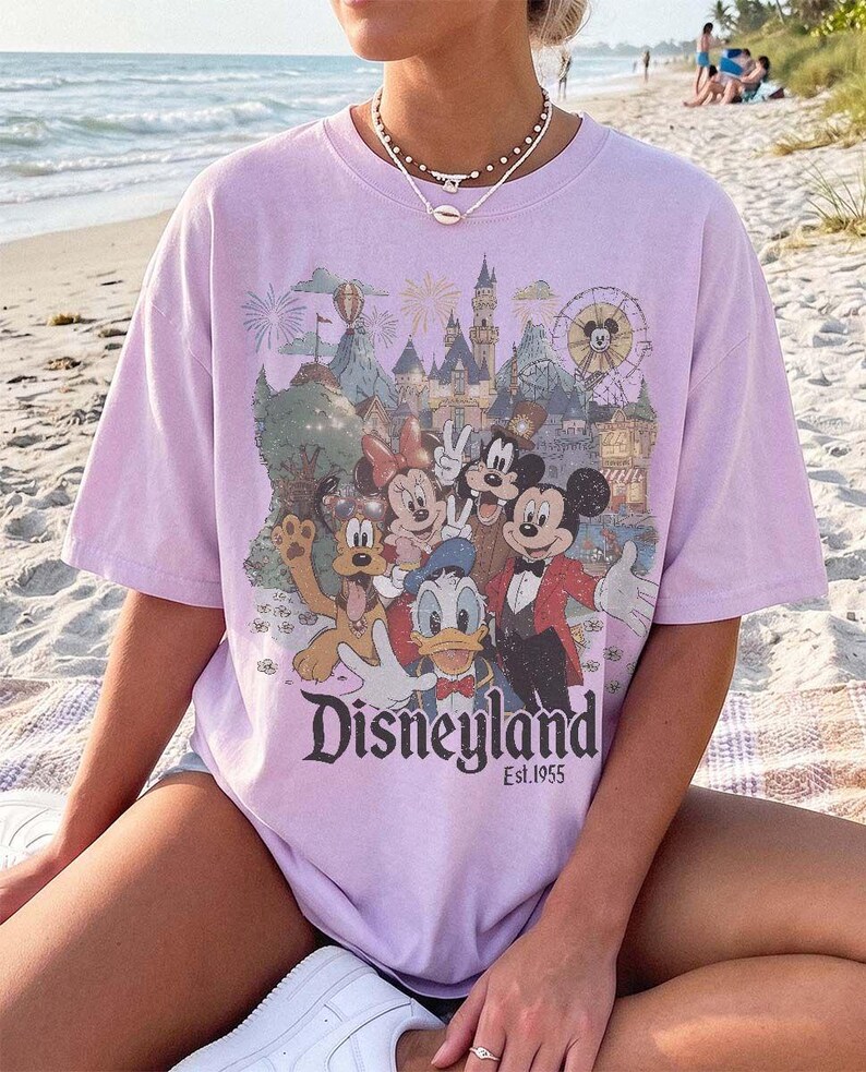 May include: A light purple short-sleeve t-shirt featuring a graphic print of Mickey Mouse, Minnie Mouse, Donald Duck, Goofy, and Pluto in front of a castle. The text Disneyland Est. 1955 is printed below the characters in a dark serif font.