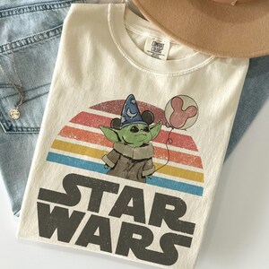 May include: Cream-colored t-shirt featuring a graphic of Grogu from Star Wars wearing a wizard hat and holding a Mickey Mouse balloon. The design is set against a retro rainbow backdrop with the words "STAR WARS" in bold letters. The shirt is folded.