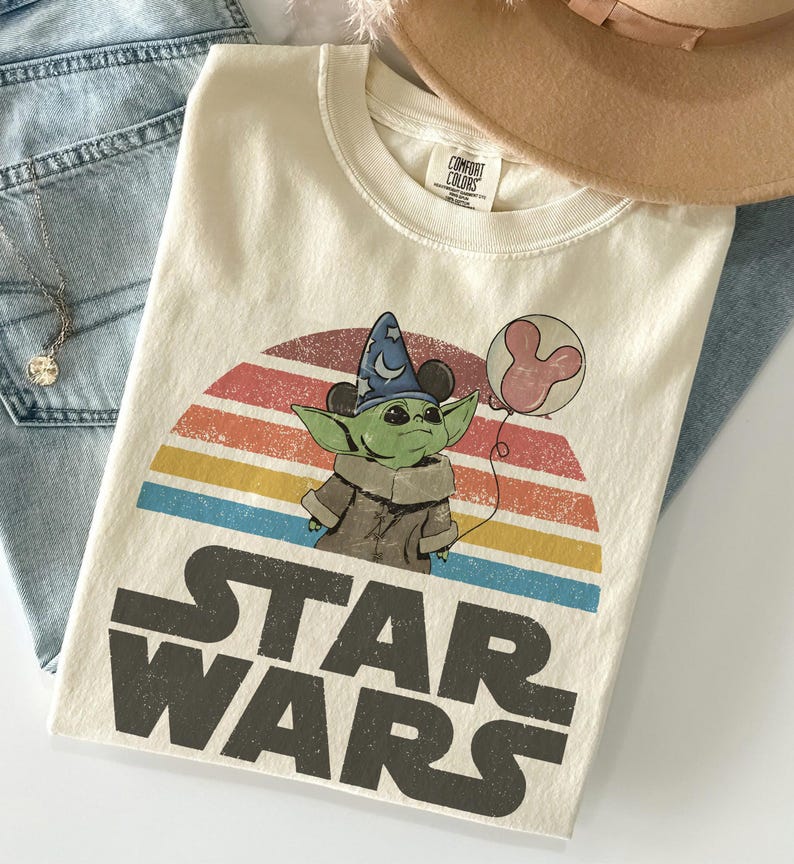 May include: Cream-colored t-shirt featuring a graphic of Grogu from Star Wars wearing a wizard hat and holding a Mickey Mouse balloon. The design is set against a retro rainbow backdrop with the words "STAR WARS" in bold letters. The shirt is folded.