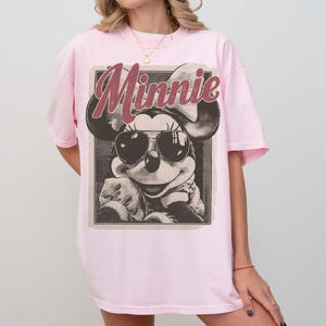 May include: A light pink t-shirt featuring a graphic print of Minnie Mouse wearing sunglasses. The word Minnie is printed in a large, distressed red font above the character. The design is enclosed in a rectangular frame with a vintage aesthetic.