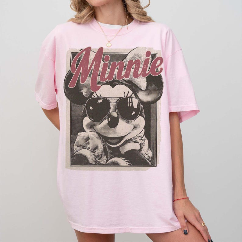 May include: A light pink t-shirt featuring a graphic print of Minnie Mouse wearing sunglasses. The word Minnie is printed in a large, distressed red font above the character. The design is enclosed in a rectangular frame with a vintage aesthetic.