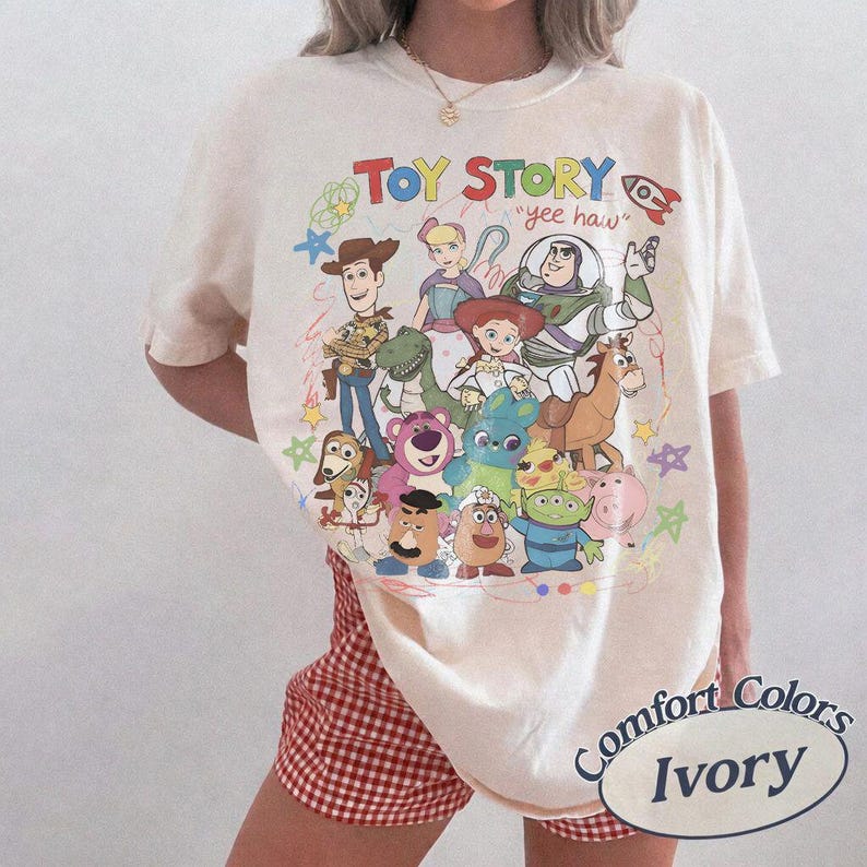 May include: An ivory t-shirt featuring a colorful graphic print of Toy Story characters including Woody, Buzz Lightyear, Jessie, and others. The text "TOY STORY" and "yee haw" appear above the characters, surrounded by small hand-drawn style stars.