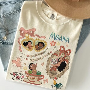 Puede incluir: Camiseta color crema con la palabra "Moana" en escritura azul, e ilustraciones de personajes y elementos de la pel&iacute;cula animada. El dise&ntilde;o incluye retratos enmarcados, una taza de t&eacute; y un cerdo. Un sombrero beige y unos vaqueros azules est&aacute;n en el fondo.