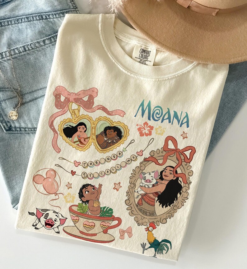 Puede incluir: Camiseta color crema con la palabra "Moana" en escritura azul, e ilustraciones de personajes y elementos de la pel&iacute;cula animada. El dise&ntilde;o incluye retratos enmarcados, una taza de t&eacute; y un cerdo. Un sombrero beige y unos vaqueros azules est&aacute;n en el fondo.