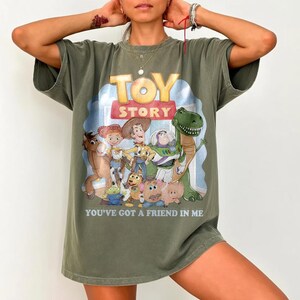 K&ouml;nnte beinhalten: Olivgr&uuml;nes T-Shirt mit einer farbenfrohen Grafik von Figuren aus dem Animationsfilm "Toy Story". Das Design enth&auml;lt die W&ouml;rter "TOY STORY" und "YOU'VE GOT A FRIEND IN ME."