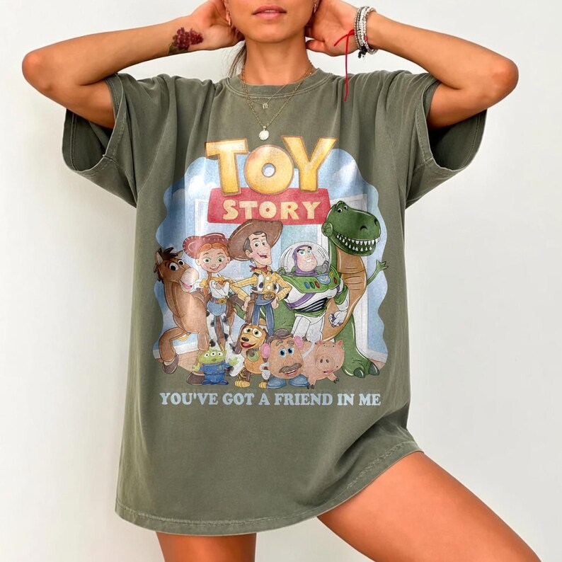 K&ouml;nnte beinhalten: Olivgr&uuml;nes T-Shirt mit einer farbenfrohen Grafik von Figuren aus dem Animationsfilm "Toy Story". Das Design enth&auml;lt die W&ouml;rter "TOY STORY" und "YOU'VE GOT A FRIEND IN ME."