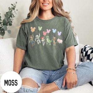K&ouml;nnte beinhalten: Moosgr&uuml;nes T-Shirt mit einer Grafik von Zeichentrickfiguren, die Mickey Mouse-f&ouml;rmige Ballons halten. Zu den Figuren geh&ouml;ren Woody, Buzz Lightyear, Jessie, Rex, Hamm und Slinky Dog. Das Shirt ist ein bequemer, l&auml;ssiger Stil.