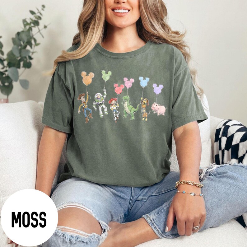 K&ouml;nnte beinhalten: Moosgr&uuml;nes T-Shirt mit einer Grafik von Zeichentrickfiguren, die Mickey Mouse-f&ouml;rmige Ballons halten. Zu den Figuren geh&ouml;ren Woody, Buzz Lightyear, Jessie, Rex, Hamm und Slinky Dog. Das Shirt ist ein bequemer, l&auml;ssiger Stil.