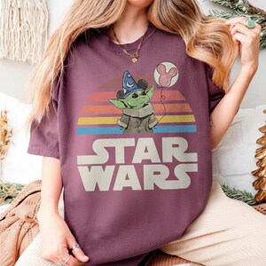 May include: A maroon Star Wars t-shirt featuring a graphic of Grogu wearing a wizard hat and holding a Mickey Mouse balloon. The design includes a retro sunset and the Star Wars logo in white.
