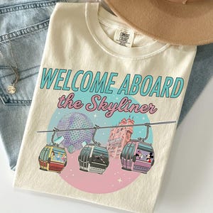 May include: Cream-colored t-shirt with the text "WELCOME ABOARD the Skyliner" in teal and pink. The graphic features a pink and blue circle with gondolas and the Spaceship Earth and Tower of Terror in the background.