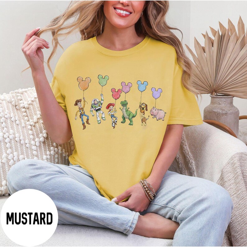 K&ouml;nnte beinhalten: Senfgelbes T-Shirt mit einer Grafik von Disney's Toy Story-Charakteren, die Mickey Mouse-f&ouml;rmige Ballons halten. Zu den Charakteren geh&ouml;ren Woody, Buzz Lightyear, Jessie, Rex, Slinky Dog und Hamm. Das Wort "MUSTARD" ist unten links aufgedruckt.