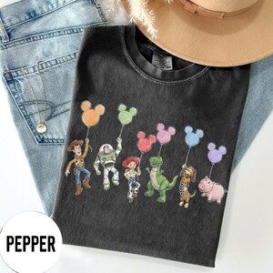 K&ouml;nnte beinhalten: Schwarzes T-Shirt mit Cartoon-Figuren aus Toy Story, die Mickey Mouse-f&ouml;rmige Ballons in verschiedenen Farben halten. Das Shirt wird mit einer Jeans und einem beigen Hut pr&auml;sentiert.