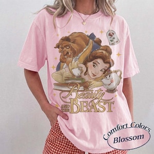 May include: A light pink short-sleeve t-shirt featuring a graphic print of Belle and the Beast from Beauty and the Beast, along with Mrs. Potts, Chip, and a rose in a glass dome. The text reads Beauty and the Beast in a gold script font.