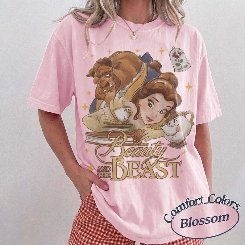 May include: A light pink short-sleeve t-shirt featuring a graphic print of Belle and the Beast from Beauty and the Beast, along with Mrs. Potts, Chip, and a rose in a glass dome. The text reads Beauty and the Beast in a gold script font.