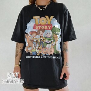K&ouml;nnte beinhalten: Schwarzes T-Shirt mit einer farbenfrohen Grafik von Charakteren aus dem Film "Toy Story". Das Design enth&auml;lt den Filmtitel und den Satz "You've Got a Friend in Me". Auf dem Shirt steht auch "Black Comfort Colors".
