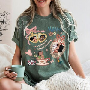 Puede incluir: Camiseta verde con la palabra "Moana" en escritura azul. La camiseta presenta ilustraciones de dibujos animados de personajes de la pel&iacute;cula, incluyendo a Moana, Hei Hei, Pua, y otros. El dise&ntilde;o incluye lazos rosas y detalles florales.