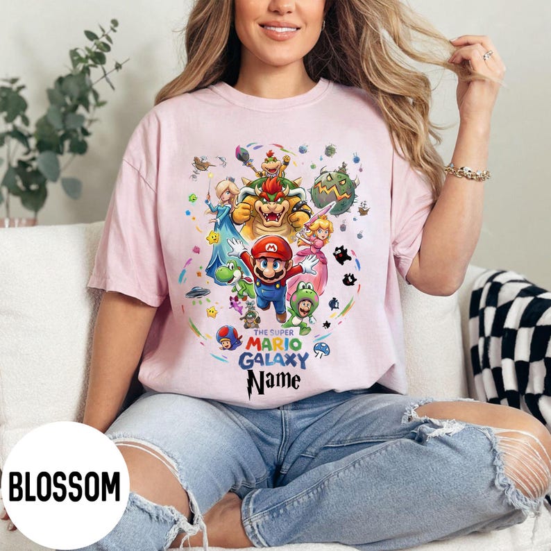Puede incluir: Camiseta rosa claro con un gr&aacute;fico colorido de Mario y otros personajes del juego Super Mario Galaxy. El texto "THE SUPER MARIO GALAXY Name" est&aacute; impreso debajo. La camiseta es de estilo casual.