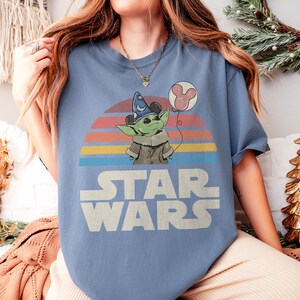 May include: A light blue Star Wars t-shirt featuring a graphic of Grogu wearing a wizard hat and holding a Mickey Mouse balloon against a retro sunset design. The words "STAR WARS" are printed in white block letters.