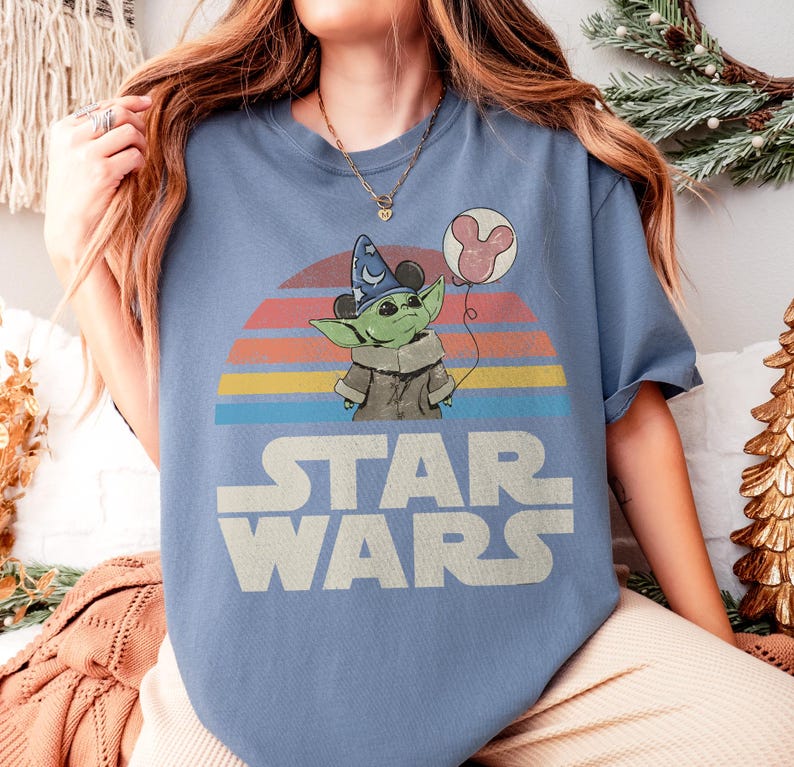 May include: A light blue Star Wars t-shirt featuring a graphic of Grogu wearing a wizard hat and holding a Mickey Mouse balloon against a retro sunset design. The words "STAR WARS" are printed in white block letters.