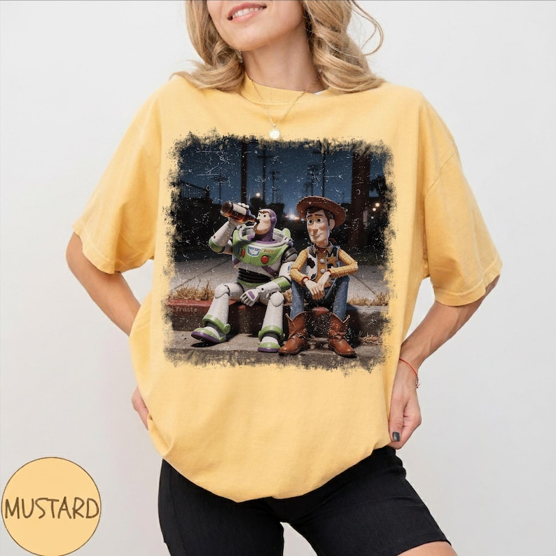 May include: A mustard yellow short-sleeve t-shirt featuring a rectangular graphic print of two animated characters sitting on a curb at night. One character is a space ranger in a white and green suit, and the other is a cowboy in a brown hat and vest.