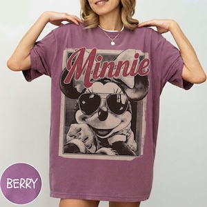May include: A berry-coloured short-sleeve t-shirt featuring a graphic print of a cartoon mouse wearing sunglasses and a bow. The text "Minnie" is printed in a large, stylized font above the character. The design has a distressed, vintage appearance.